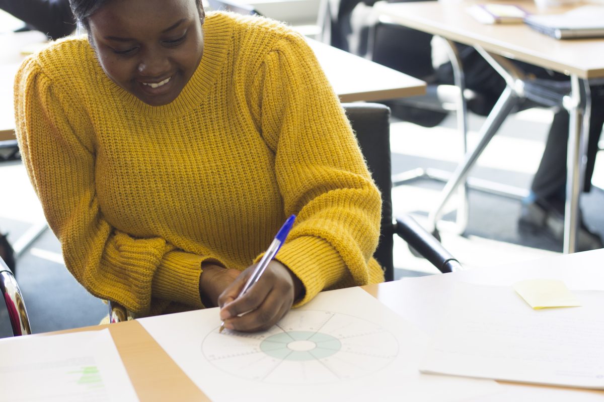 a young black women wearing a yellow sweater is writing on paper and smiling