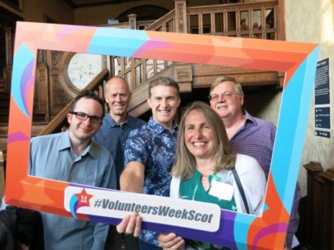 Four men and a woman are smiling and holding a picture frame with the hashtag volunteer scotland written across it 