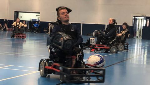 in a gymnasium a groups of people with physical disabilities are playing football in powerchairs