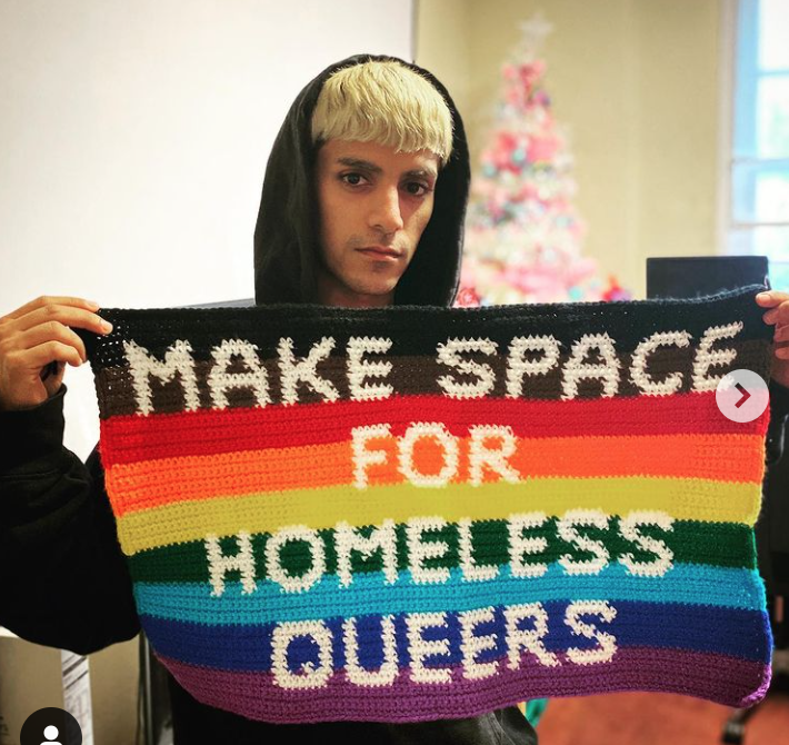 A blond man wearing a hood is holding a Pride and black Lives matter flag stating "Make Space for homeless queers"