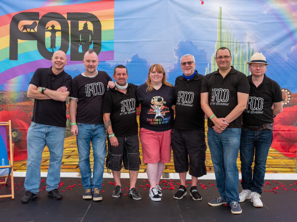 7 people wearing back t-shirt with FOD club written on it arestanding smiling at the camera. Behind them are rainbow flags