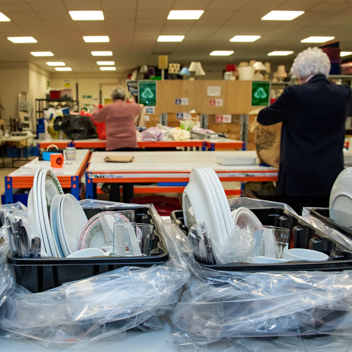 Two trays with plates and cutleries on a table. At the back of the picture are people working to put items into bags