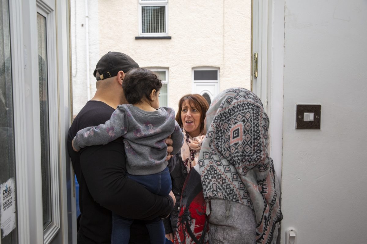 In the opening of a door are from behind a man carrying a children and a women wearing a head scarf. They are talking with a women.