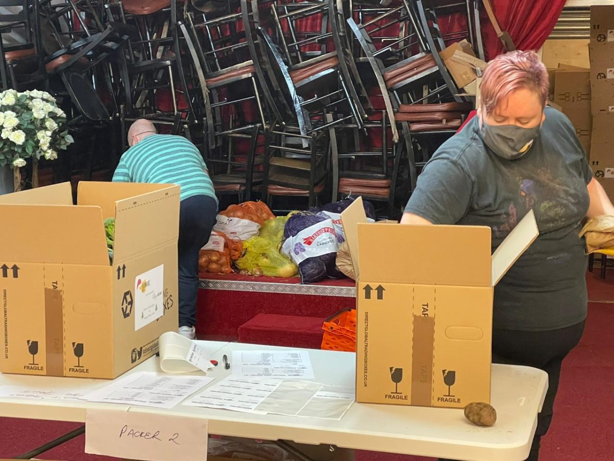 Scene of a food distribution day. At the back, nags with vegetables are put on a table. One person is working near the bags.At the front of the picture an other person is putting items in cardboard boxes. 