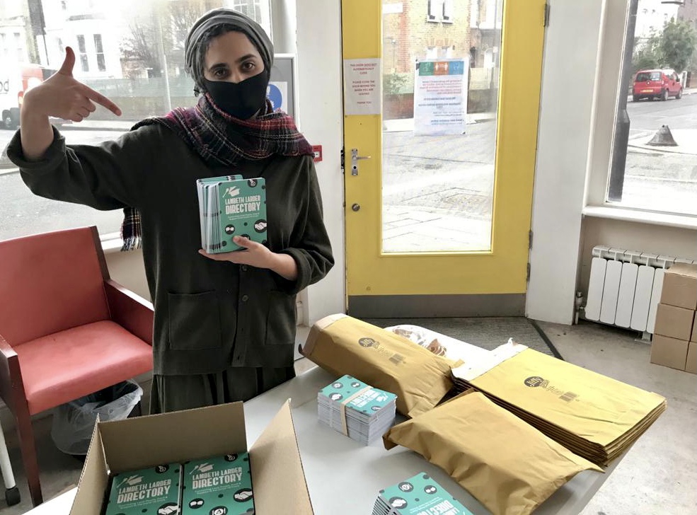 A person wearing a face mask is looking at the camera. In on hand she holds a pile of flyers with the name of the charity, and she is pointing towards them with her other hand. 