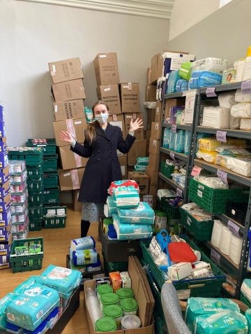 a woman is standing in a room filled with boxes, food trays, nappies, baby food and more. She is looking at the camera with her hands up and a happy look.