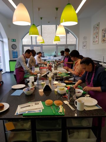 In a room, 7 people wearing aprons are gathered around a high table. They are all busy cooking. Bowls, graters and diverse ingredients are spread across the table 