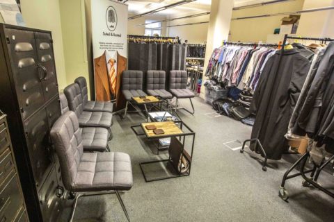 A room is filled with work clothes, costumes and shirts. At the front is a sitting area with a few chairs and tables and.a big banner with the name of the charity "suited and booted"