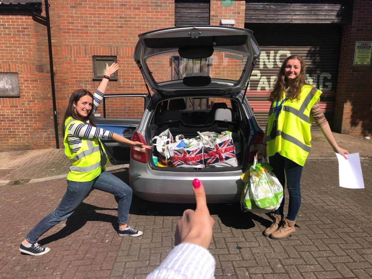 A car parked with the boot open and filled with groceries bag. On the sides two individuals wearing yellow are smilling and looking at the camera. At the front is a thumbs up