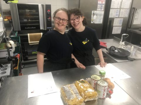 Two young people wearing black t-shirt branded with the yellow submarine logo are posing and smiling to the camera. They are standing behind a counter in a commercial kitchen.