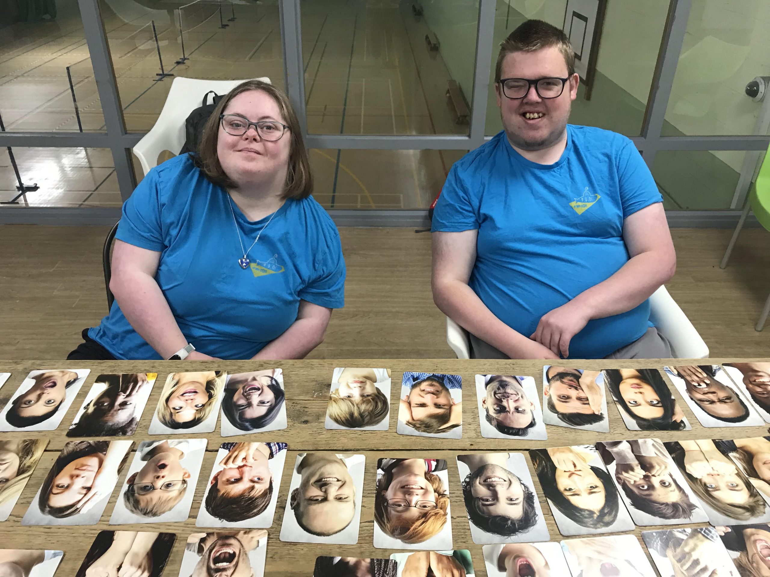 two people wearing blue t-shirt (the yellow submarine branded t shirts) are sitting behind a table. On the table they have cards with printed faces on them