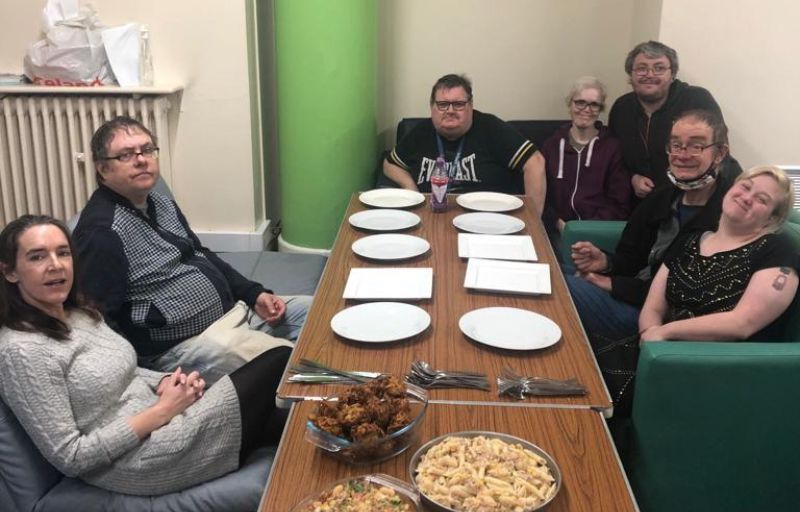 7 people are sitting around a table, looking at the camera and smiling. Empty plates are on the table with food ready to be served.