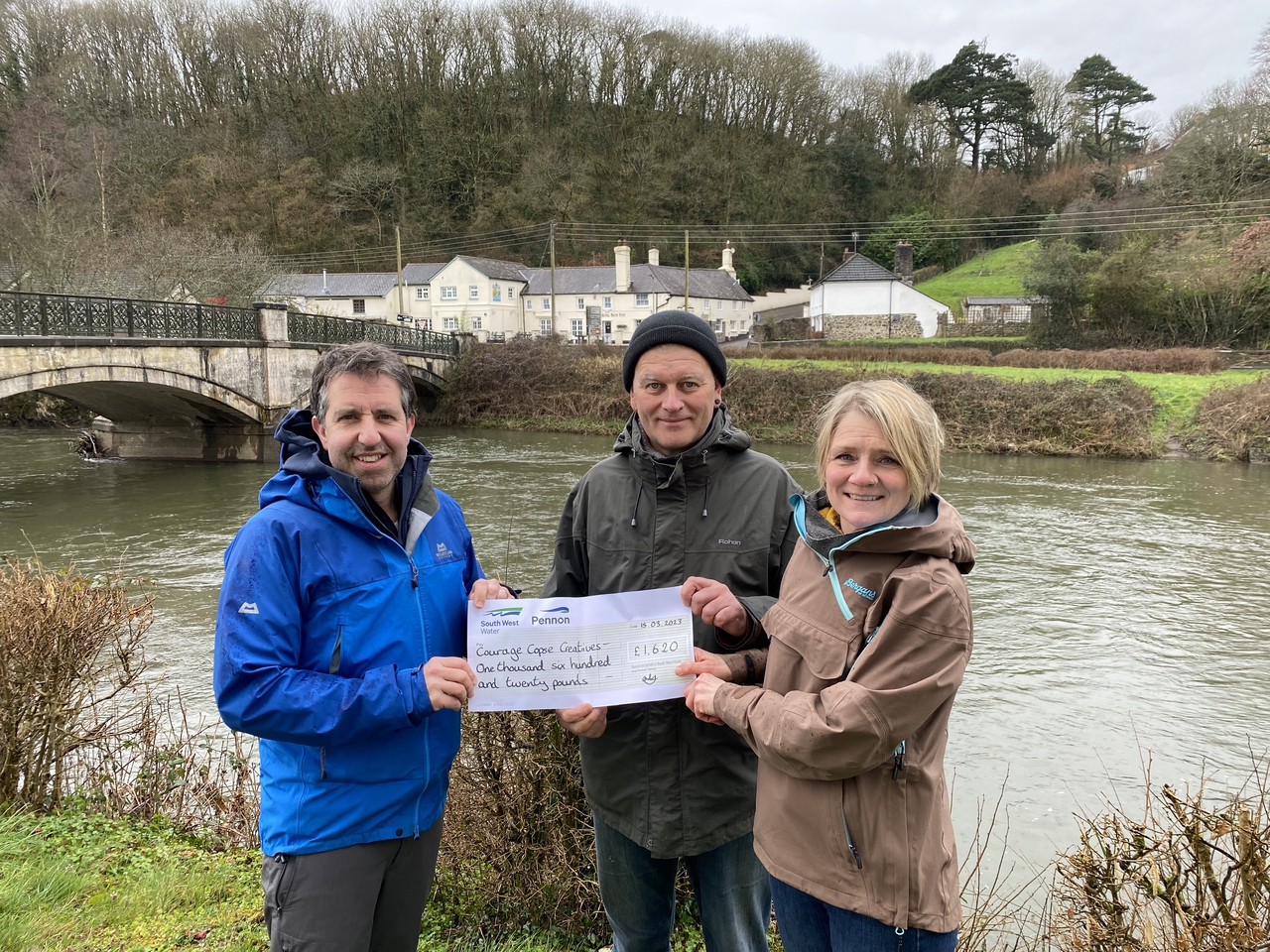 a river in the background. Three people are standing at the front. They are holding a large fake cheque and smiling at the camera